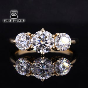 Puede incluir: Un anillo de oro de tres piedras con tres diamantes redondos grandes y transparentes. El anillo está sobre un fondo negro y se refleja en la superficie.