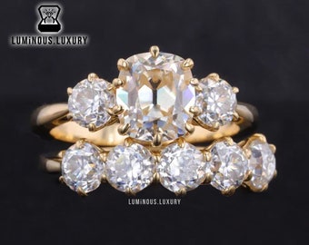 Puede incluir: Un anillo de compromiso de oro con una piedra central grande de corte cojín y piedras redondas más pequeñas en la banda. El anillo está sobre un fondo negro.