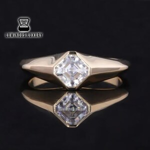 Peut inclure: Une bague en or avec une pierre précieuse carrée transparente sertie au centre. La bague a une bande épaisse avec un dessus légèrement arrondi.