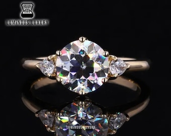 May include: A gold engagement ring with a large round center stone and two smaller pear-shaped stones on either side. The ring is set on a black background.