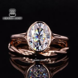 Puede incluir: Un anillo de compromiso de oro rosa con un gran diamante de forma ovalada. El anillo está engastado con una banda delicada y una banda de boda a juego con un diseño trenzado.