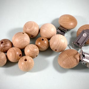 May include: A collection of wooden beads and pacifier clips. The beads are various sizes and have a natural wood grain finish. The pacifier clips have a silver metal clip attached to a wooden disc. The items are arranged on a light blue surface.