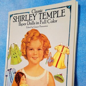 May include: A vintage paper doll book featuring Shirley Temple. The cover shows a color illustration of Shirley Temple holding a doll, with various paper doll outfits and accessories surrounding her. The book title reads "Classic Shirley Temple Paper Dolls in Full Color" and is edited by Grayce Piemonte.