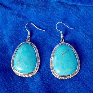 May include: A pair of turquoise teardrop earrings with silver-colored metal trim and ear hooks. The turquoise stones have a marbled appearance and are set in a silver-colored metal frame with a rope-like border. The earrings are displayed on a blue surface.