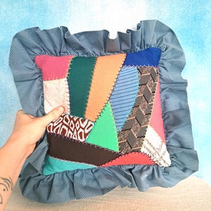 May include: A patchwork pillow with a blue ruffled border. The pillow is made from various fabrics in shades of blue, green, pink, orange, and brown. The fabrics are sewn together in a random pattern.