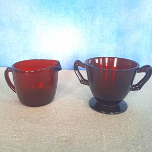 May include: A set of two vintage red glass creamer and sugar bowls. The creamer has a handle and a pouring spout. The sugar bowl has two handles and a pedestal base.
