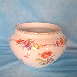 May include: A white ceramic bowl featuring a rounded shape and a wide opening. The exterior is decorated with a colorful floral pattern of orange, pink, yellow, and purple flowers. A thin gold-colored decorative trim lines the top rim of the bowl.