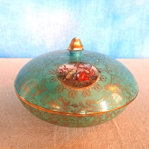 May include: A teal ceramic trinket box with a gold lid and gold trim. The lid features a painted scene of two figures in a garden setting.