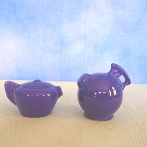 May include: Two small, matching, purple ceramic pieces. One is a teapot with a lid and handle. The other is a round creamer with a handle. Both pieces are glossy and sit on a beige surface against a blue background.