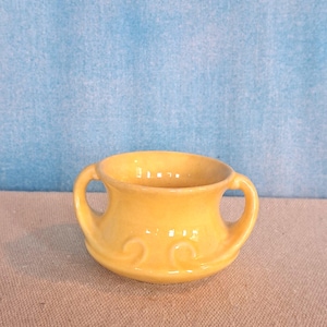 May include: A small, yellow ceramic bowl with two handles. The bowl has a rounded shape with decorative swirls near the base. The background is a light blue color.