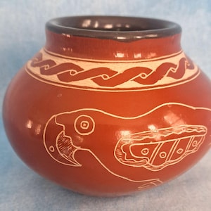 May include: A small, round, terracotta-colored ceramic pot with a dark brown rim. The exterior features a white etched design of a parrot on the side and a continuous wave pattern encircling the neck of the vessel, creating a decorative contrast.
