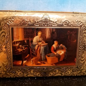 May include: A gold metal box with an ornate design and a picture of a woman sewing with a baby in a cradle. The box has a hinged lid and is decorated with a floral pattern.