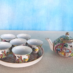 May include: A white porcelain tea set with a teapot and six cups. The set features a colorful floral and figure design. The cups are arranged on a white tray with a floral design.