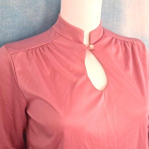 May include: A dusty rose colored blouse featuring a high mandarin collar, a small keyhole cutout at the center neckline, and a single button closure. The garment has gathered fabric details at the shoulder seams and three-quarter length sleeves.