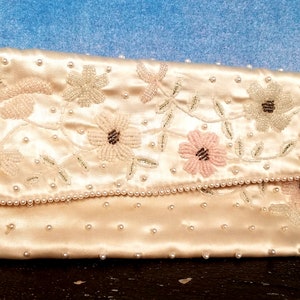 May include: A white satin clutch purse with a beaded floral design. The purse is decorated with pearls and has a flap closure.