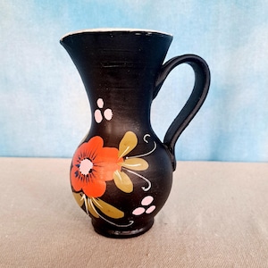 May include: A black ceramic pitcher with a handle, featuring a hand-painted floral design. The flower is primarily orange with a pink center and green and yellow leaves. White and pink dots accent the design. The pitcher has a white rim.