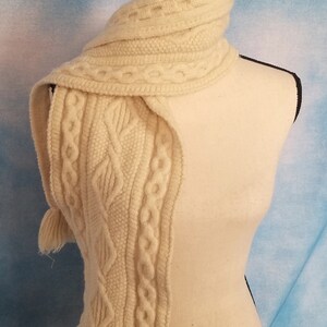 May include: A white knitted scarf with a cable knit pattern and fringe. The scarf is displayed on a white mannequin.