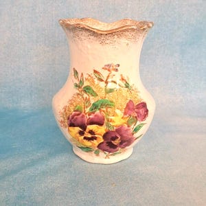 May include: A white ceramic vase with a scalloped rim, adorned with a floral design featuring purple and yellow pansies. The vase has a gold trim and a bulbous shape, standing on a light blue background.