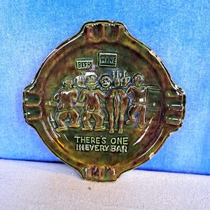 May include: Green ceramic ashtray with a raised design of four figures at a bar with the words "Beer" and "Wine" above them. The text "There's one in every bar" is written below the figures.