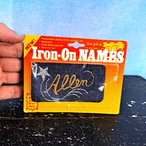 May include: A yellow and orange package with the text "Iron-On NAMES" and "three fish inc."  The package contains a blue denim patch with the name "Allen" embroidered in gold thread.