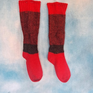May include: A pair of knee-high socks with a color block design. The socks feature red cuffs and toes, with a dark gray band separating the red sections from the dark red main body of the socks. The socks are displayed on a light blue background.