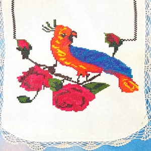 May include: A white embroidered fabric with a colorful bird and roses. The bird is orange, blue, and yellow. The roses are pink and green.