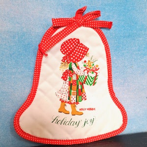 May include: A white fabric Christmas stocking with red polka dots around the edge. The stocking features a girl in a red and white polka dot dress holding a green and white gift box. The text "HOLLY HOBBIE" and "holiday joy" is printed on the stocking.