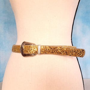 May include: A gold glitter belt with a silver buckle and a matching silver loop. The belt is wrapped around a mannequin. The belt is approximately 1.5 inches wide.