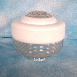 May include: A glass light fixture shade featuring a clear, ribbed lower section and a smooth, opaque white upper band. The top of the shade is crowned with a circular, textured glass insert displaying a raised, repeating floral pattern.