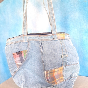 May include: Light blue denim tote bag with a zipper closure and denim straps. The bag features colorful plaid fabric patches and a small pocket with a label. The bag is held up against a light blue background.