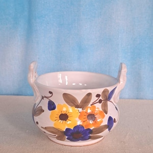 May include: White ceramic bowl with two handles, adorned with hand-painted floral designs in yellow, orange, and blue. Brown leaves and stems accent the flowers. The bowl is suitable for decorative or functional use.