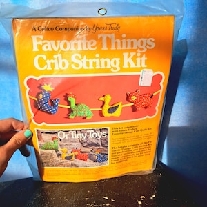 May include: A yellow and orange package with a picture of a crib string kit featuring a variety of colorful animals. The package says "Favorite Things Crib String Kit" and "Or Tiny Toys".