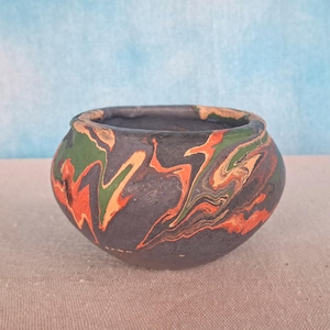 May include: A small, round ceramic bowl with a dark gray base and a marbled design in orange, green, and tan. The bowl has a wide opening and a slightly textured surface. The background is a light blue.