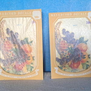 May include: Two vintage yellow and brown paper cards with the text "MEYERCORD DECALS" and "10c" printed on them. Each card features a colorful image of a cornucopia overflowing with fruit and grapes. The text "EASY TO APPLY ALL YOU USE IS WATER" is printed at the bottom of each card.