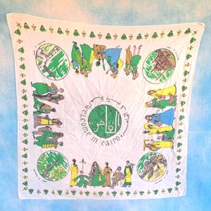 May include: White square scarf with a green border and illustrations. The center features text in English and Arabic, with a circular design. The scarf has drawings of people and landmarks, with green, yellow, and blue accents.