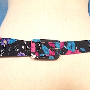 May include: A black fabric belt with a floral print in shades of pink, blue, and purple. The belt has a black plastic buckle.