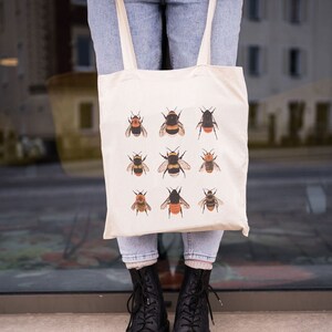 Op de afbeelding: Een witte tote bag met een ontwerp van negen hommels in verschillende poses. De tas heeft twee lange banden en wordt gedragen door iemand die blauwe jeans en zwarte laarzen draagt.