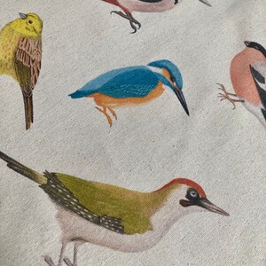 Puede incluir: Primer plano de una tela con un patr&oacute;n de cinco p&aacute;jaros diferentes. Los p&aacute;jaros son un pinz&oacute;n amarillo, un p&aacute;jaro carpintero verde, un mart&iacute;n pescador azul, un p&aacute;jaro carpintero blanco y negro y un p&aacute;jaro rosa y negro. La tela es de color beige claro.