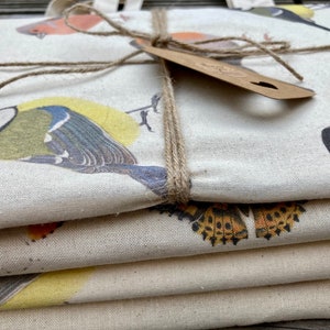 Puede incluir: Una pila de cuatro bolsas de tela con un dise&ntilde;o de estampado de p&aacute;jaros. Las bolsas est&aacute;n hechas de lona de color natural y presentan una variedad de p&aacute;jaros, incluyendo un p&aacute;jaro azul, un petirrojo y un jilguero. Las bolsas est&aacute;n atadas juntas con un cordel.
