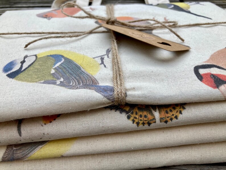 Puede incluir: Una pila de cuatro bolsas de tela con un dise&ntilde;o de estampado de p&aacute;jaros. Las bolsas est&aacute;n hechas de lona de color natural y presentan una variedad de p&aacute;jaros, incluyendo un p&aacute;jaro azul, un petirrojo y un jilguero. Las bolsas est&aacute;n atadas juntas con un cordel.
