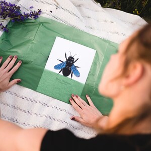 以下が含まれることがあります： 青い大工蜂の印刷されたイラストのクローズアップ。白いカードに「Blaue Holzbiene」というテキストが書かれており、緑色の薄紙の背景があります。