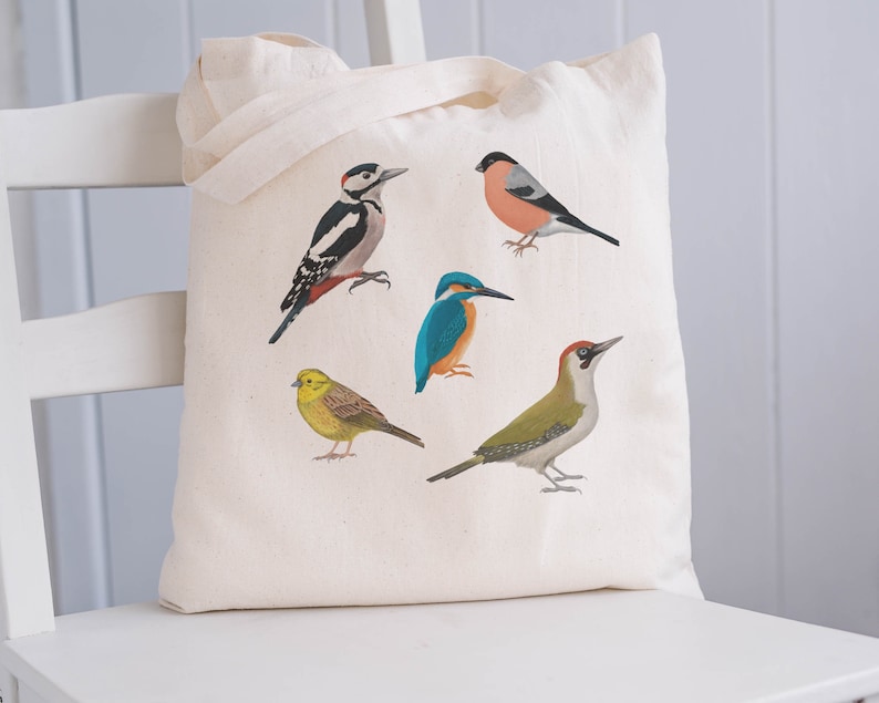 Puede incluir: Una bolsa de lona blanca con cinco ilustraciones de diferentes p&aacute;jaros. Los p&aacute;jaros son un p&aacute;jaro carpintero, un petirrojo, un mart&iacute;n pescador, un verder&oacute;n y un jilguero.