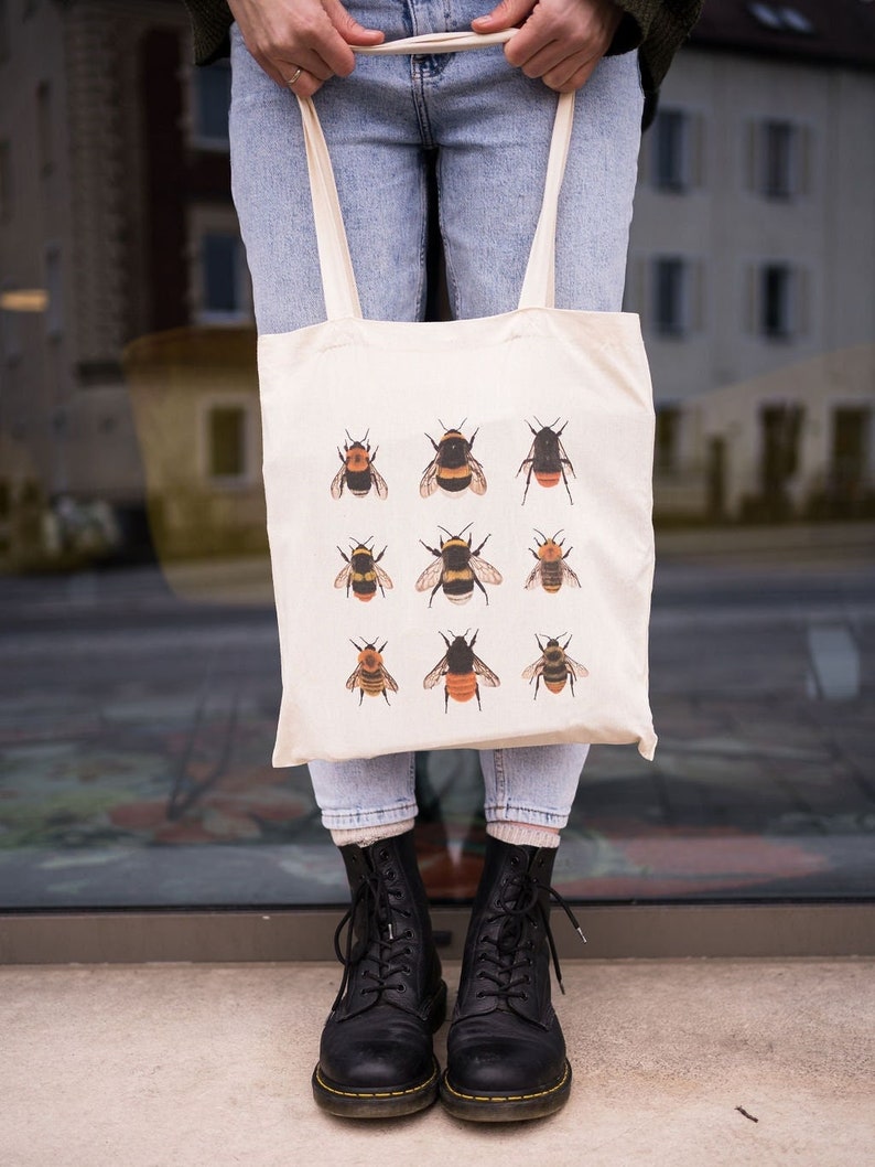 Op de afbeelding: Een witte tote bag met een ontwerp van negen hommels in verschillende poses. De tas heeft twee lange banden en wordt gedragen door iemand die blauwe jeans en zwarte laarzen draagt.