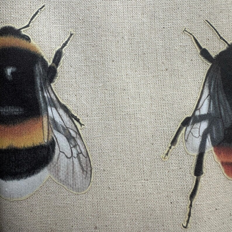Op de afbeelding: Twee hommels met zwarte en gele strepen en doorschijnende vleugels zijn genaaid op een beige canvas stof.