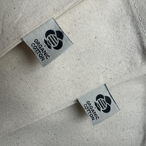 Op de afbeelding: Twee witte stoffen tassen met zwarte labels waarop "Organic Cotton" staat en die een gestileerd zwart bloemenontwerp hebben.