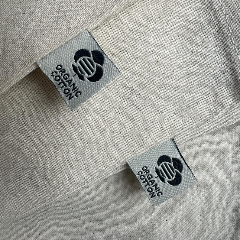 Op de afbeelding: Twee witte stoffen tassen met zwarte labels waarop "Organic Cotton" staat en die een gestileerd zwart bloemenontwerp hebben.