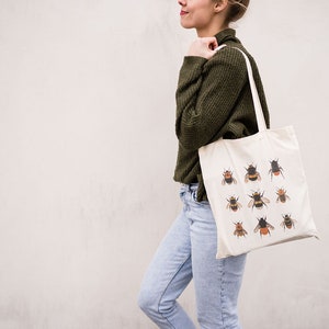 Op de afbeelding: Een vrouw draagt een witte tote bag met een ontwerp van hommels. De tas heeft bruine hengsels en is gemaakt van canvas.