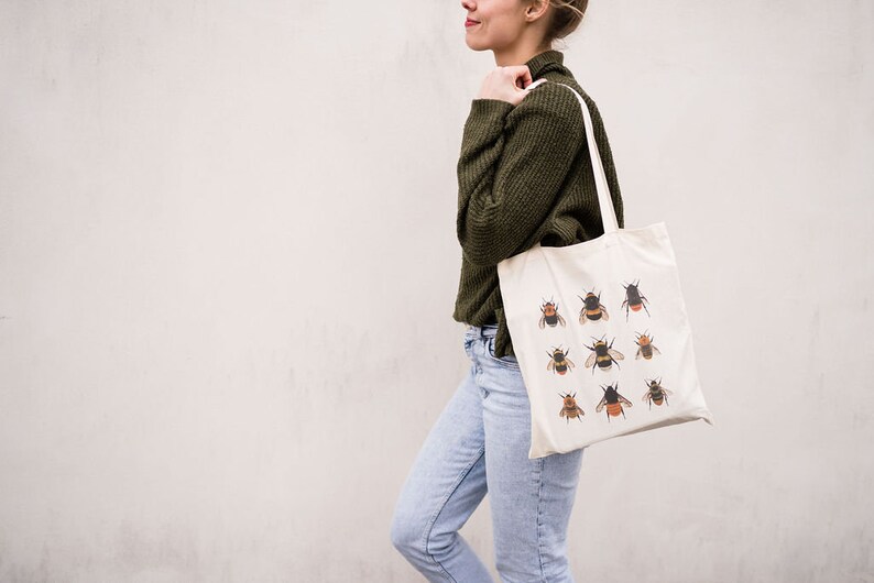 Op de afbeelding: Een vrouw draagt een witte tote bag met een ontwerp van hommels. De tas heeft bruine hengsels en is gemaakt van canvas.