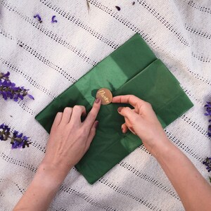 Puede incluir: Una persona est&aacute; sellando un sobre verde con un sello de cera dorado que dice "Danke". El sobre est&aacute; sobre una manta blanca de ganchillo con flores moradas.