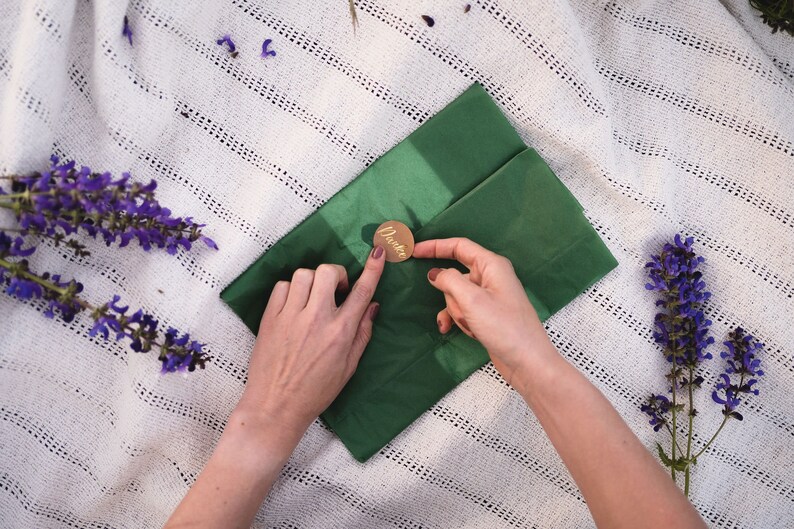 Puede incluir: Una persona est&aacute; sellando un sobre verde con un sello de cera dorado que dice "Danke". El sobre est&aacute; sobre una manta blanca de ganchillo con flores moradas.